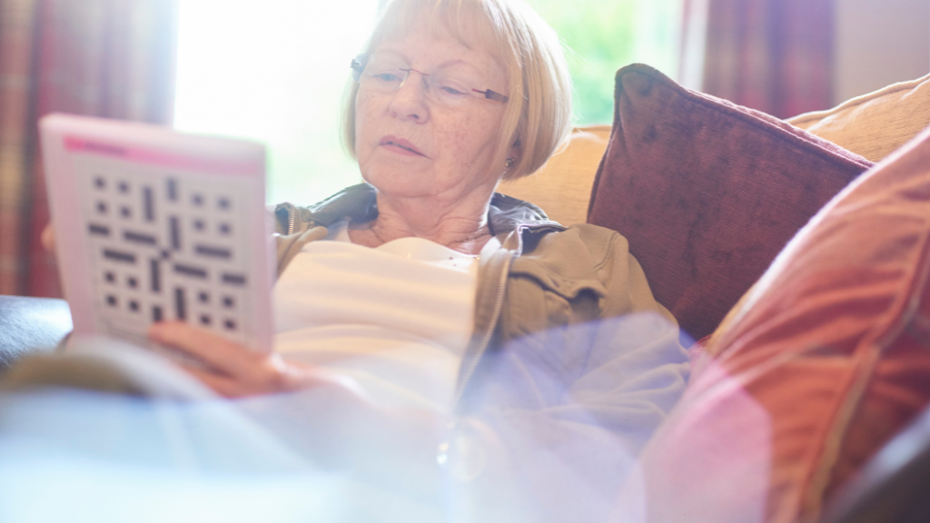 An older woman works on a crossword puzzle while sitting on the couch.
