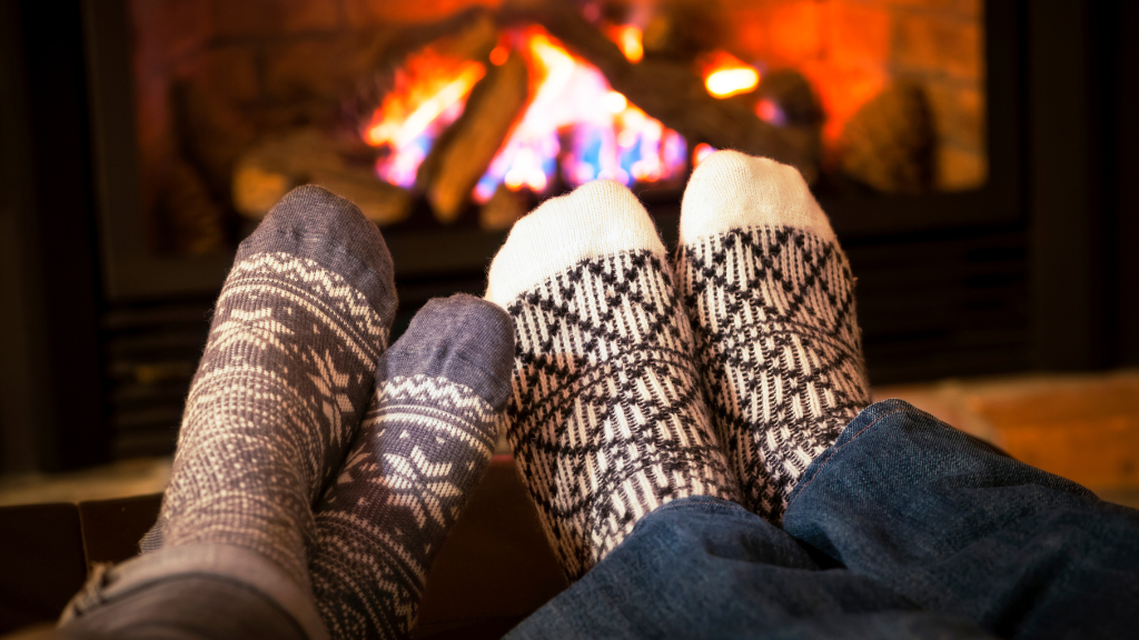 Two sets of feet wearing winter-themed socks in front of a roaring fireplace.
