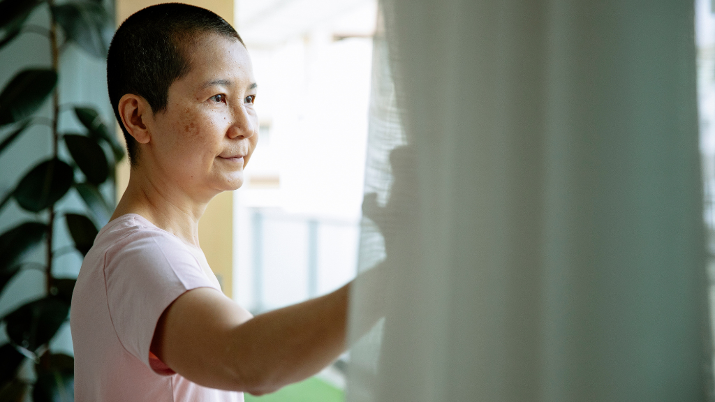 A middle-aged Asian woman with buzzed hair smiles hopefully while pulling back the curtain of her window.